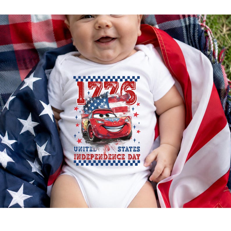 A baby wearing a white onesie with a graphic of a red car and the text "1776 United States Independence Day" is lying on a blanket with an American flag.