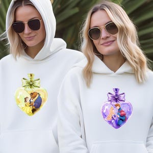 Puede incluir: Dos sudaderas con capucha blancas con un dise&ntilde;o de botella en forma de coraz&oacute;n. Una botella es amarilla con Bella y la Bestia de La Bella y la Bestia. La otra botella es morada con un pr&iacute;ncipe y una princesa.
