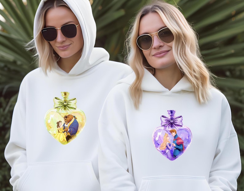 Puede incluir: Dos sudaderas con capucha blancas con un dise&ntilde;o de botella en forma de coraz&oacute;n. Una botella es amarilla con Bella y la Bestia de La Bella y la Bestia. La otra botella es morada con un pr&iacute;ncipe y una princesa.