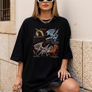 Peut inclure: T-shirt noir ample avec quatre illustrations de dragons, dont les noms sont "Andarna", "Sgaepi" et "Tain". Les dragons sont repr&eacute;sent&eacute;s en or, argent et bleu. Le t-shirt a des manches courtes.