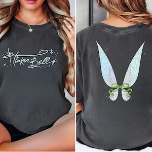 May include: Dark gray t-shirt featuring the text "Tinker Bell" in a whimsical script on the front. The back of the shirt displays a colorful fairy wing design with green ribbon accents. A comfortable, casual style.