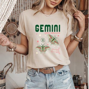 Może przedstawiać: Beżowa koszulka z zielonym napisem "GEMINI". Poniżej znajduje się napis "ADAPTABLE, CURIOUS, & CLEVER". Projekt zawiera ilustracje motyli i r&oacute;żowy układ gwiazd.