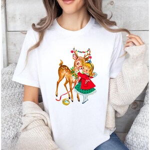 Op de afbeelding: Wit T-shirt met een vintage kerstillustratie. De afbeelding toont een hert versierd met ornamenten en een meisje in een rode jas en groene jurk. Een feestelijk ontwerp in retrostijl, perfect voor de feestdagen.