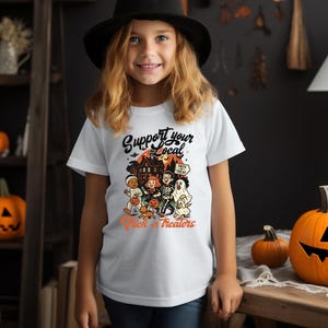A young girl wearing a white t-shirt with a Halloween-themed graphic and text, standing in a room decorated for Halloween.