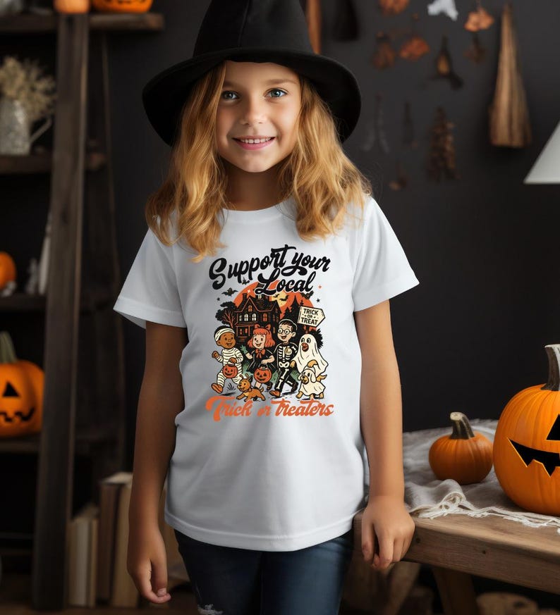 A young girl wearing a white t-shirt with a Halloween-themed graphic and text, standing in a room decorated for Halloween.