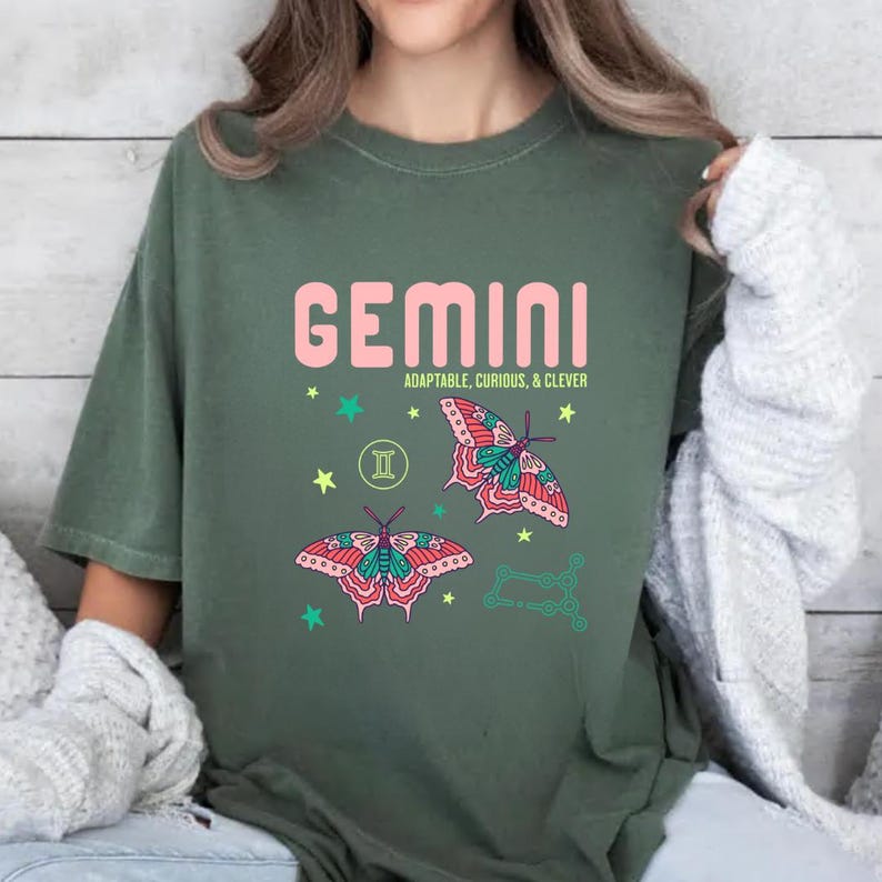 Może przedstawiać: Zielona koszulka z napisem "GEMINI" w r&oacute;żowych literach. Poniżej słowa znajdują się słowa "ADAPTABLE, CURIOUS, & CLEVER". Koszulka przedstawia dwie ilustracje motyli z r&oacute;żowymi, turkusowymi i czerwonymi skrzydłami, a także grafikę gwiazd i konstelacji.