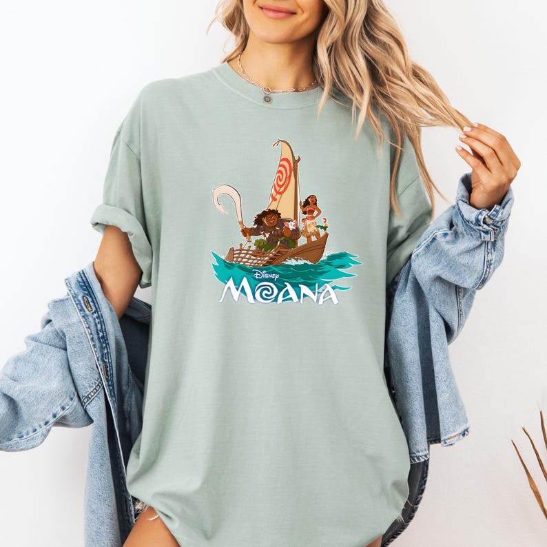 May include: A light blue t-shirt with a graphic of Maui, Moana, and Pua sailing on a boat. The text "Disney Moana" is printed below the image.