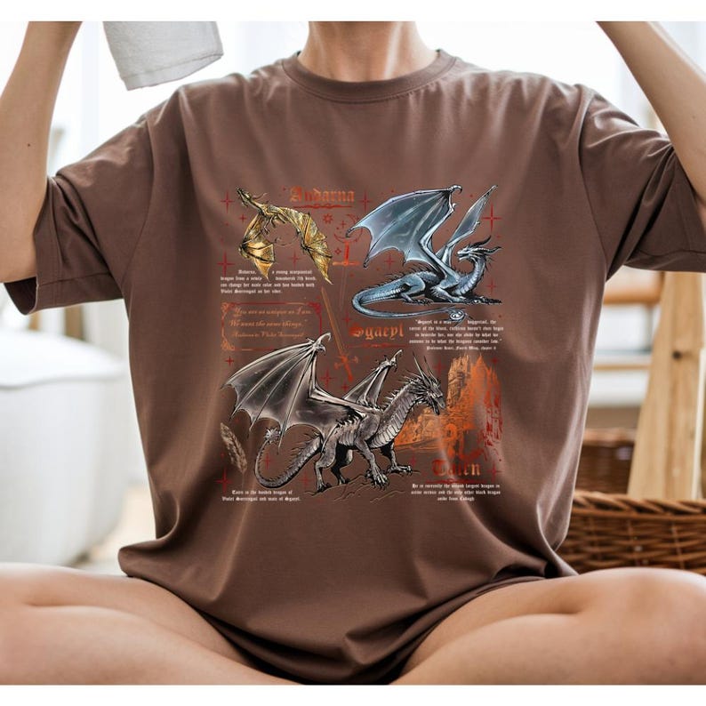 Peut inclure: T-shirt marron avec des illustrations de dragons et du texte. Le t-shirt a un col rond et des manches courtes. Les illustrations de dragons sont en or, bleu et argent, avec du texte de style vintage.
