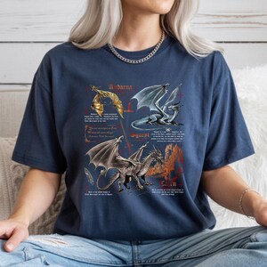 Peut inclure: T-shirt bleu marine avec trois illustrations de dragons et du texte. Les dragons sont repr&eacute;sent&eacute;s en or, argent et bleu, avec un ch&acirc;teau en arri&egrave;re-plan. Le texte comprend des noms et des descriptions de dragons.