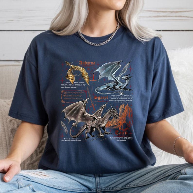 Peut inclure: T-shirt bleu marine avec trois illustrations de dragons et du texte. Les dragons sont repr&eacute;sent&eacute;s en or, argent et bleu, avec un ch&acirc;teau en arri&egrave;re-plan. Le texte comprend des noms et des descriptions de dragons.