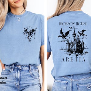 May include: Light blue t-shirt with a black dragon and moon graphic on the front and a castle with dragons on the back. The text "RIORSON HOUSE ARETIA" is printed on the back. The shirt is made of a soft material.