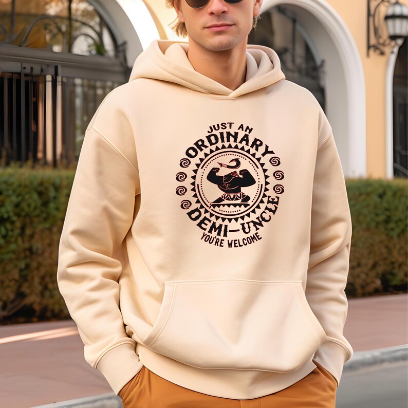 May include: A cream-coloured hoodie with a front pocket and a drawstring hood. The hoodie displays a circular graphic with the text "JUST AN ORDINARY DEMI-UNCLE YOU'RE WELCOME" in a brown and black design. The graphic includes an illustration of a muscular figure.