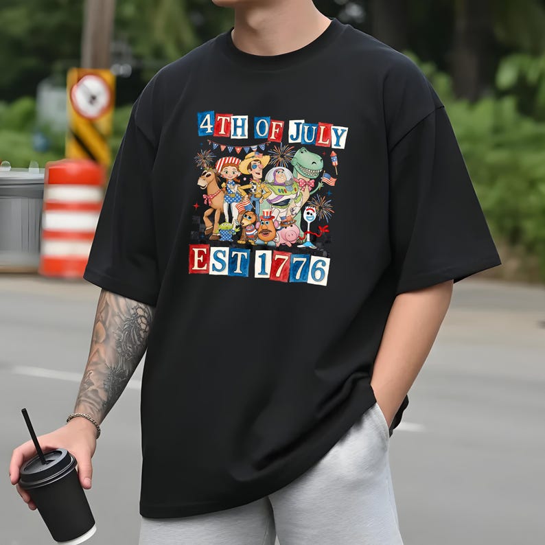 A person wearing a black t-shirt with a colorful graphic design depicting various cartoon characters and fireworks, celebrating the 4th of July.