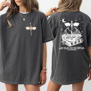 a woman wearing a gray t-shirt with a graphic design on the back. The design features a bat, a crescent moon, and a book, along with the text "I AM THE SKY AND THE POWERS OF EVERY STORM THAT HAS EVER BEEN, I AM INFINITE".