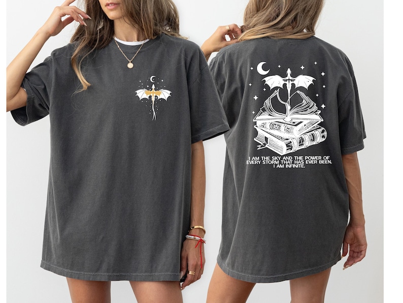 a woman wearing a gray t-shirt with a graphic design on the back. The design features a bat, a crescent moon, and a book, along with the text "I AM THE SKY AND THE POWERS OF EVERY STORM THAT HAS EVER BEEN, I AM INFINITE".