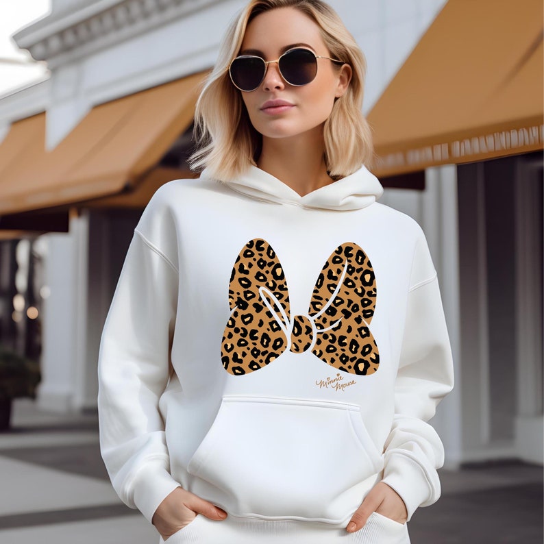 May include: A white hoodie featuring a large bow design with a leopard print pattern. The bow is centered on the front of the hoodie, with a small signature below. The hoodie has a kangaroo pocket and a drawstring hood.