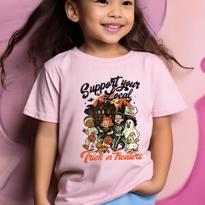 A young girl wearing a pink t-shirt with a Halloween-themed graphic and text, smiling at the camera.