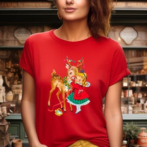 Op de afbeelding: Rood T-shirt met een vintage illustratie van een meisje dat een hert omhelst, versierd met kerstverlichting. Het meisje draagt een rode jas en groene jurk. Onderaan staat een klein mandje.