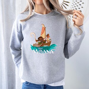 May include: A light gray sweatshirt featuring a graphic of characters from the Disney movie Moana sailing on a boat. The design includes the movie title in white and a stylized ocean scene.