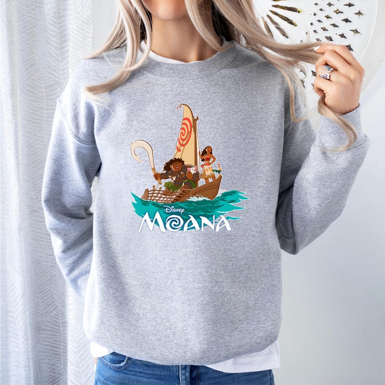 May include: A light gray sweatshirt featuring a graphic of characters from the Disney movie Moana sailing on a boat. The design includes the movie title in white and a stylized ocean scene.