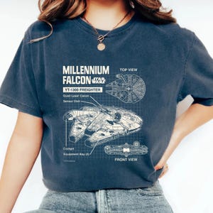 May include: A navy blue t-shirt with a white graphic of the Millennium Falcon spaceship from Star Wars. The graphic includes a top and front view of the ship with details like the quad laser canon, sensor dish, cockpit, and equipment bay.