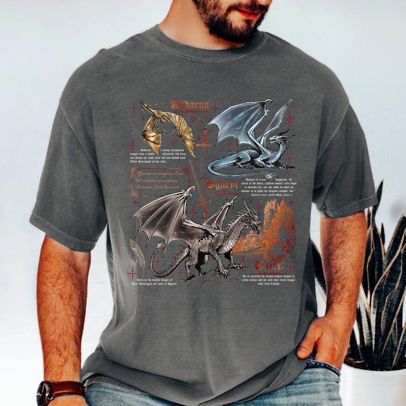 Peut inclure: T-shirt gris avec trois illustrations de dragons et du texte. Les dragons sont repr&eacute;sent&eacute;s en or, bleu et gris, avec des ailes et des &eacute;cailles d&eacute;taill&eacute;es. Le texte est de style m&eacute;di&eacute;val, sur fond rouge d&eacute;lav&eacute;.