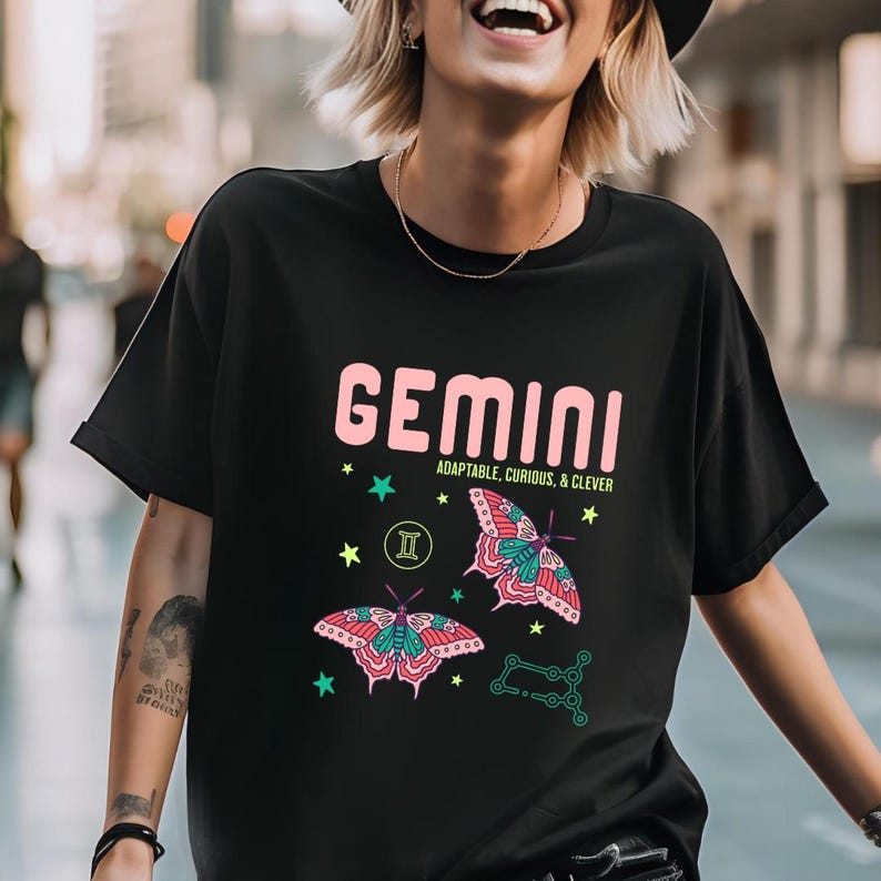 Może przedstawiać: Czarna koszulka z wzorem znaku zodiaku Bliźnięta. Wz&oacute;r zawiera r&oacute;żowy napis "GEMINI", frazę "ADAPTABLE, CURIOUS, & CLEVER", dwie ilustracje motyli oraz grafikę gwiazd i konstelacji.