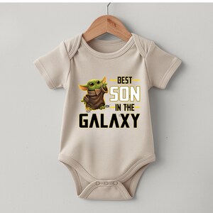 Pu&ograve; includere: Un body da bambino beige con un personaggio alieno verde e la scritta "BEST SON in the GALAXY" in nero e giallo. Il body &egrave; appeso a una gruccia di legno.