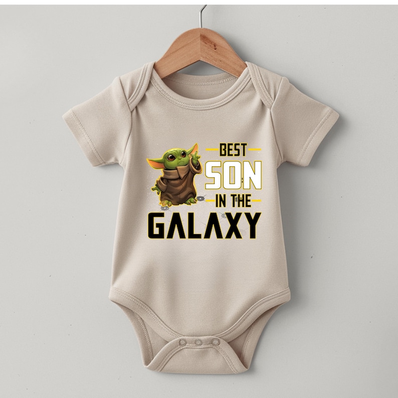 Pu&ograve; includere: Un body da bambino beige con un personaggio alieno verde e la scritta "BEST SON in the GALAXY" in nero e giallo. Il body &egrave; appeso a una gruccia di legno.