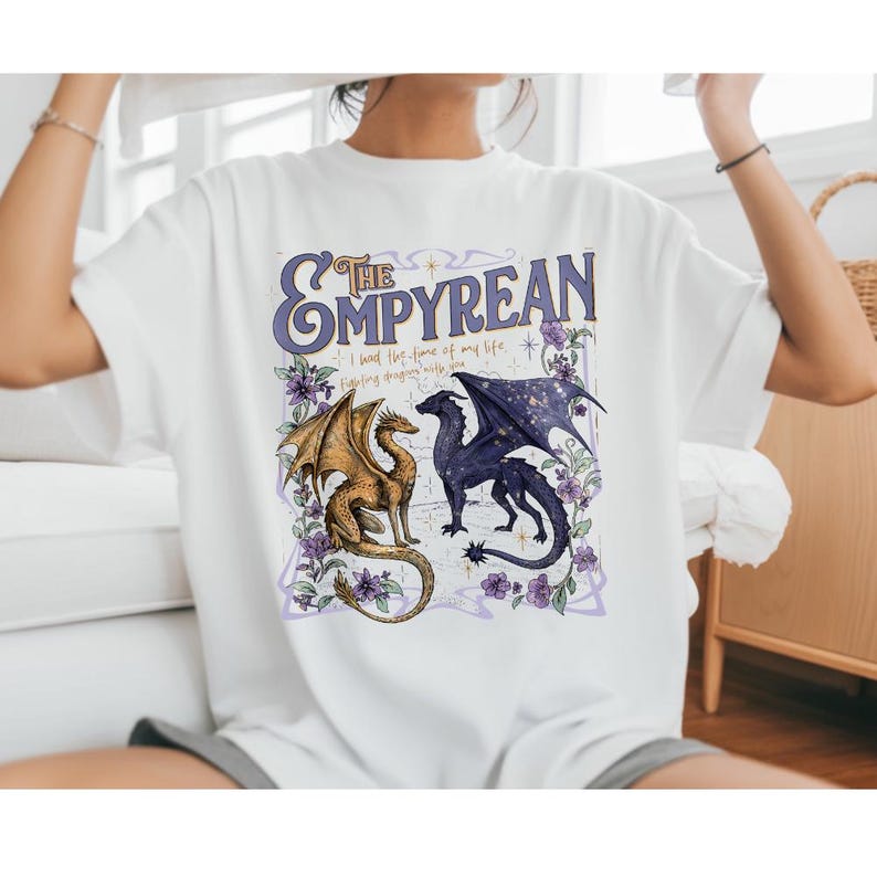 Pode incluir: Camiseta branca com um design gr&aacute;fico com o texto "The Empyrean" em roxo, e dois drag&otilde;es. Um drag&atilde;o &eacute; dourado, e o outro &eacute; azul escuro com um design de asas estreladas. A camiseta tamb&eacute;m tem o texto "I had the time of my life... fighting dragons with you."