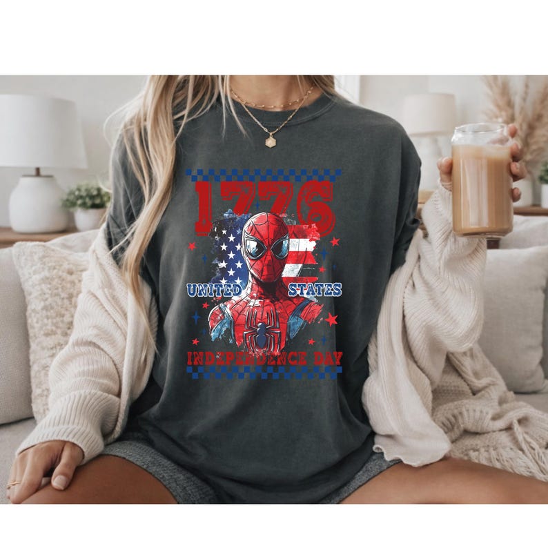 May include: Dark grey t-shirt featuring a graphic of Spider-Man with an American flag design. The text "1776", "United States", and "Independence Day" are displayed in red and blue.