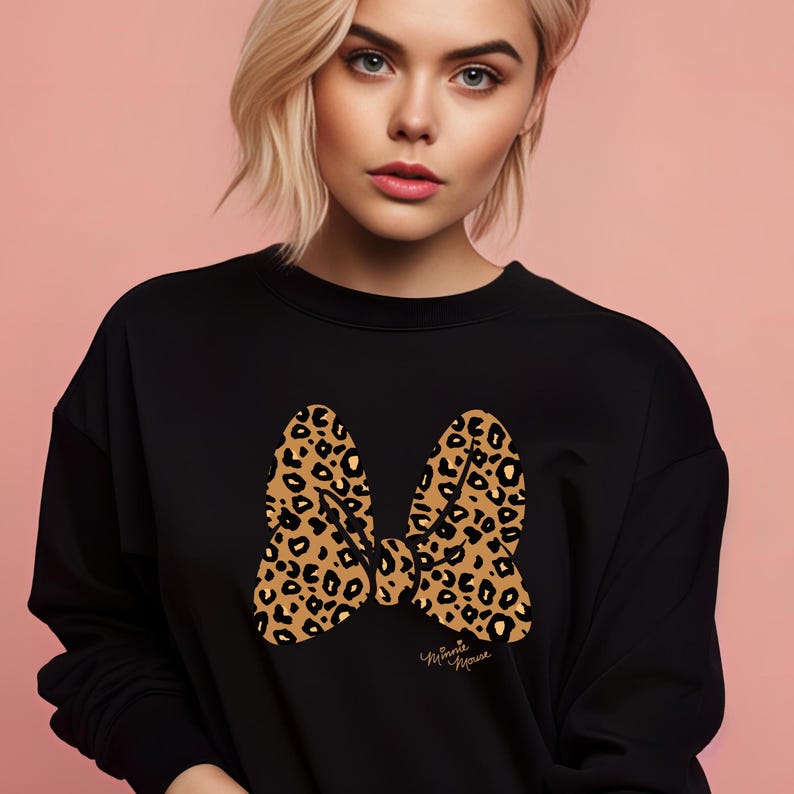 May include: Black sweatshirt featuring a large leopard print bow design. The bow is in shades of brown and tan with black spots. The words "Minnie Mouse" are written in cursive below the bow.