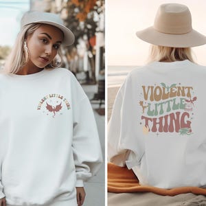 May include: White sweatshirt with a colorful graphic print that says "Violent Little Thing" with a dragon and other small details. The sweatshirt is shown on a person wearing a hat.