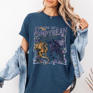 Pode incluir: Camiseta azul marinho com um design gr&aacute;fico. O design inclui o texto "The Empyrean" em roxo, ilustra&ccedil;&otilde;es de dois drag&otilde;es e detalhes florais. A camiseta tem gola redonda e mangas curtas.