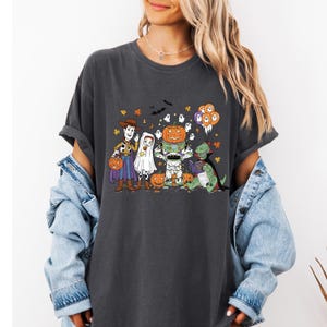 A woman wearing a black t-shirt with a Halloween-themed graphic design featuring characters such as Woody, a ghost, a skeleton, a pumpkin, a dragon, and a bride and groom.