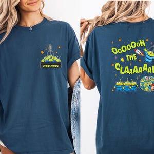 May include: Navy blue t-shirt featuring a cartoon design of the aliens from the movie Toy Story. The front of the shirt has a claw machine graphic with the text "EST.1995". The back of the shirt has the text "OooOOoH the CLAAAAAW".