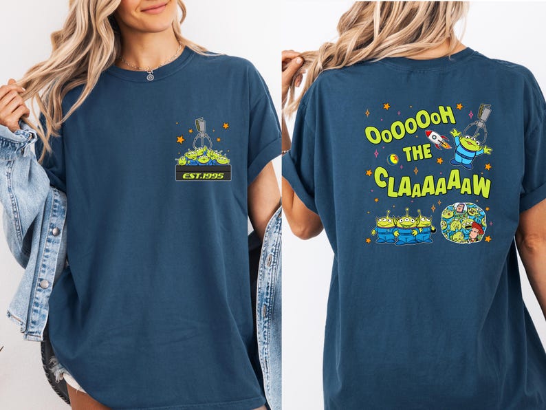 May include: Navy blue t-shirt featuring a cartoon design of the aliens from the movie Toy Story. The front of the shirt has a claw machine graphic with the text "EST.1995". The back of the shirt has the text "OooOOoH the CLAAAAAW".