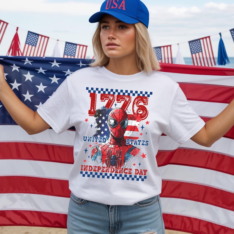 May include: White t-shirt featuring a graphic of Spider-Man with the text "1776 United States Independence Day." The design incorporates the American flag colors: red, white, and blue. A blue baseball cap with "USA" is also visible.