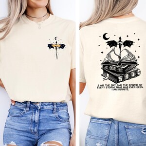 a woman wearing a white t-shirt with a graphic design on the back. The design features a dragon and a crescent moon, along with the text "I am the sky and the power of every storm that ever been, I am infinite".
