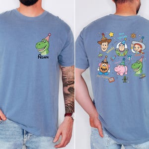 Puede incluir: Camiseta azul con un dinosaurio de dibujos animados con un gorro de fiesta y el nombre "Adam". La parte trasera de la camiseta muestra personajes de Toy Story con gorros de fiesta, como Woody, Buzz Lightyear y Jessie.