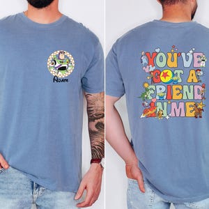 May include: A blue t-shirt featuring a Buzz Lightyear graphic on the front with the name "Adam" below. The back of the shirt displays the phrase "You've Got a Friend in Me" with various Toy Story characters in a colorful design.