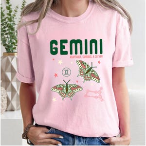 Może przedstawiać: Jasnor&oacute;żowa koszulka z napisem "GEMINI" w ciemnozielonych literach. Poniżej słowa znajdują się słowa "ADAPTABLE, CURIOUS, & CLEVER". Koszulka ma r&oacute;wnież dwie ilustracje motyli i symbol zodiaku Bliźnięta.