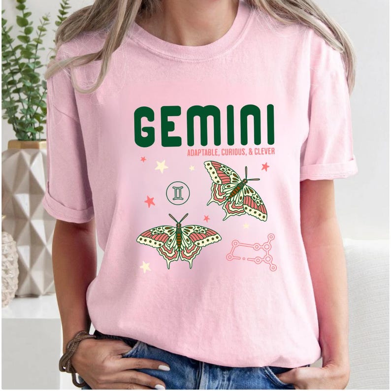 Może przedstawiać: Jasnor&oacute;żowa koszulka z napisem "GEMINI" w ciemnozielonych literach. Poniżej słowa znajdują się słowa "ADAPTABLE, CURIOUS, & CLEVER". Koszulka ma r&oacute;wnież dwie ilustracje motyli i symbol zodiaku Bliźnięta.