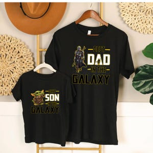 Pu&ograve; includere: Due magliette nere con disegni abbinati. Una maglietta dice "BEST DAD IN THE GALAXY" con una grafica di Mandaloriano. L'altra dice "BEST SON IN THE GALAXY" con una grafica di Grogu. Entrambe le magliette sono appese a una gruccia di legno.