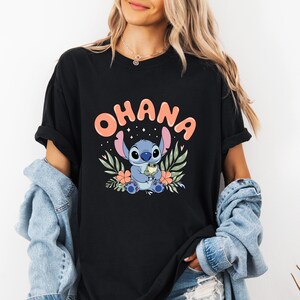 May include: Black t-shirt featuring the word "OHANA" in coral letters above a blue cartoon character holding a small green creature. The design includes floral and leaf accents in green and orange.