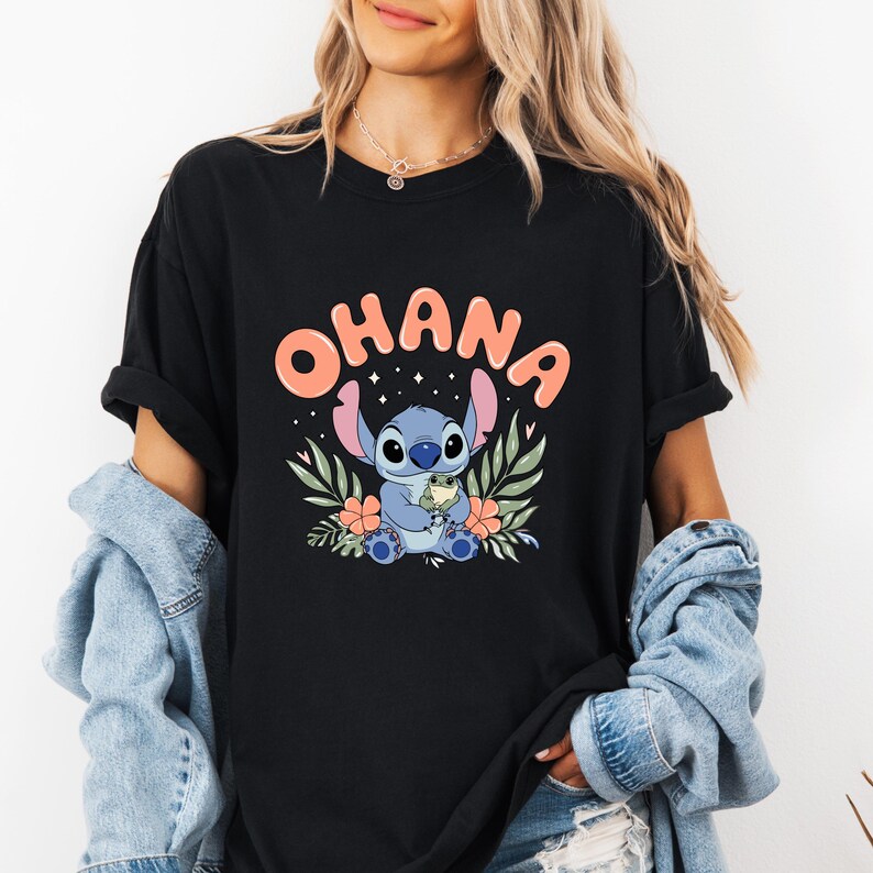 May include: Black t-shirt featuring the word "OHANA" in coral letters above a blue cartoon character holding a small green creature. The design includes floral and leaf accents in green and orange.