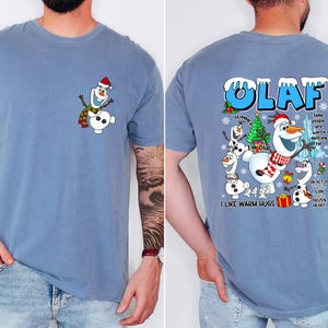 Op de afbeelding: Lichtblauw T-shirt met een cartoon van Olaf de sneeuwman met een hoed en sjaal. De achterkant van het shirt heeft een groter Olaf-ontwerp met de tekst "OLAF" en "I like warm hugs."