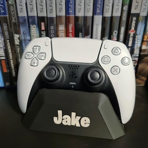 May include: A white and black PlayStation 5 controller on a black stand with the name "Jake". The controller is positioned in front of a shelf filled with various video game cases.