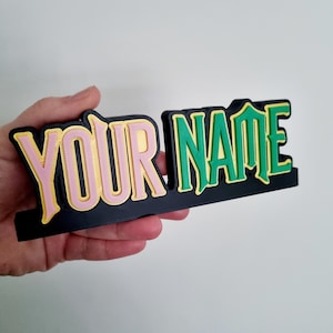 May include: A black 3D printed nameplate with the words "YOUR NAME" in pink, green, and gold lettering.
