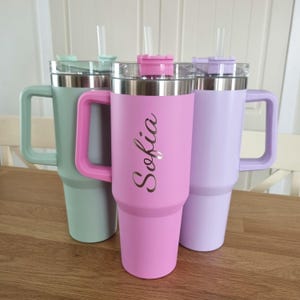 May include: Three pastel-coloured insulated tumblers: mint green, pink, and lavender. Each has a handle, a clear lid, and a stainless steel rim. The pink tumbler is personalised with the name "Sofia". Clear straws are inserted into the lids.